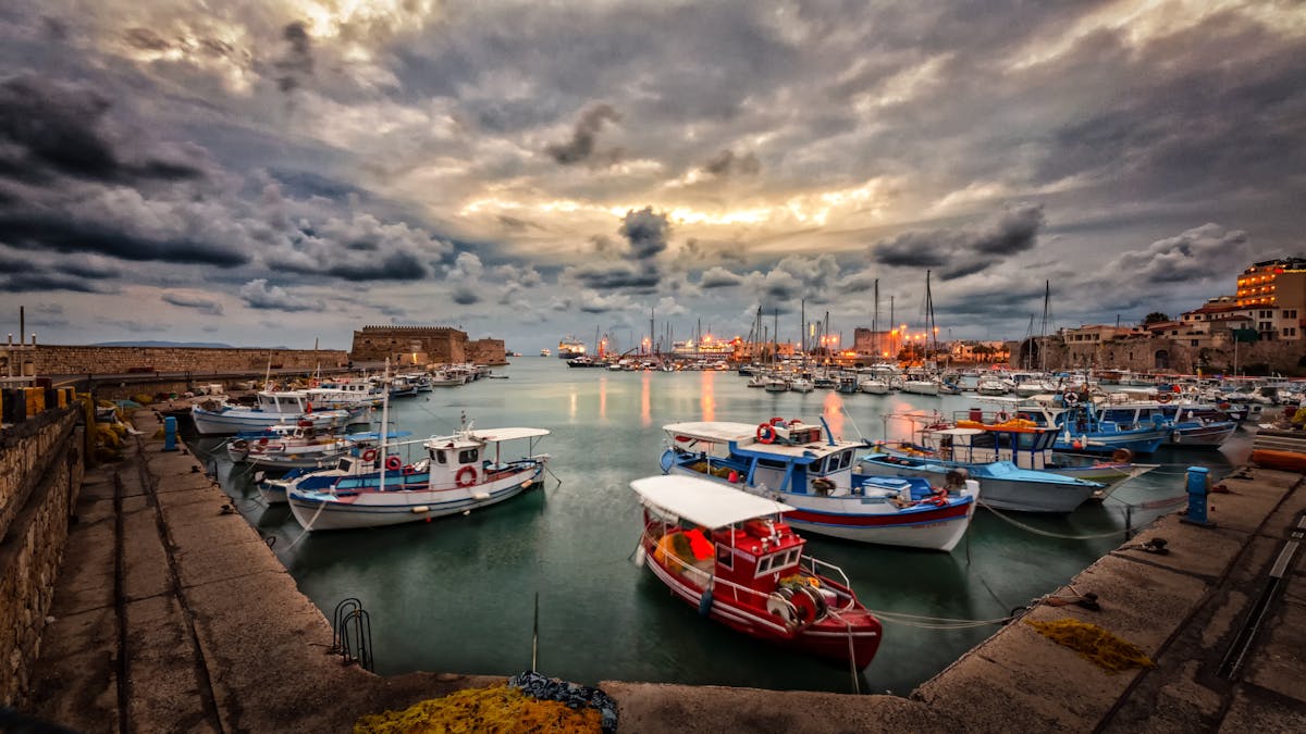 A cluster of small fishing boats moored inside a stone-walled Mediterranean harbour under heavy clouds in low dawn light