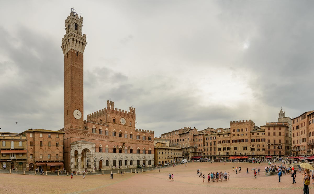 A Tuscan piazza with a tall medieval brick tower and surrounding stone palazzi under an overcast sky, the kind of civic square around which a sculpture scene tends to cluster