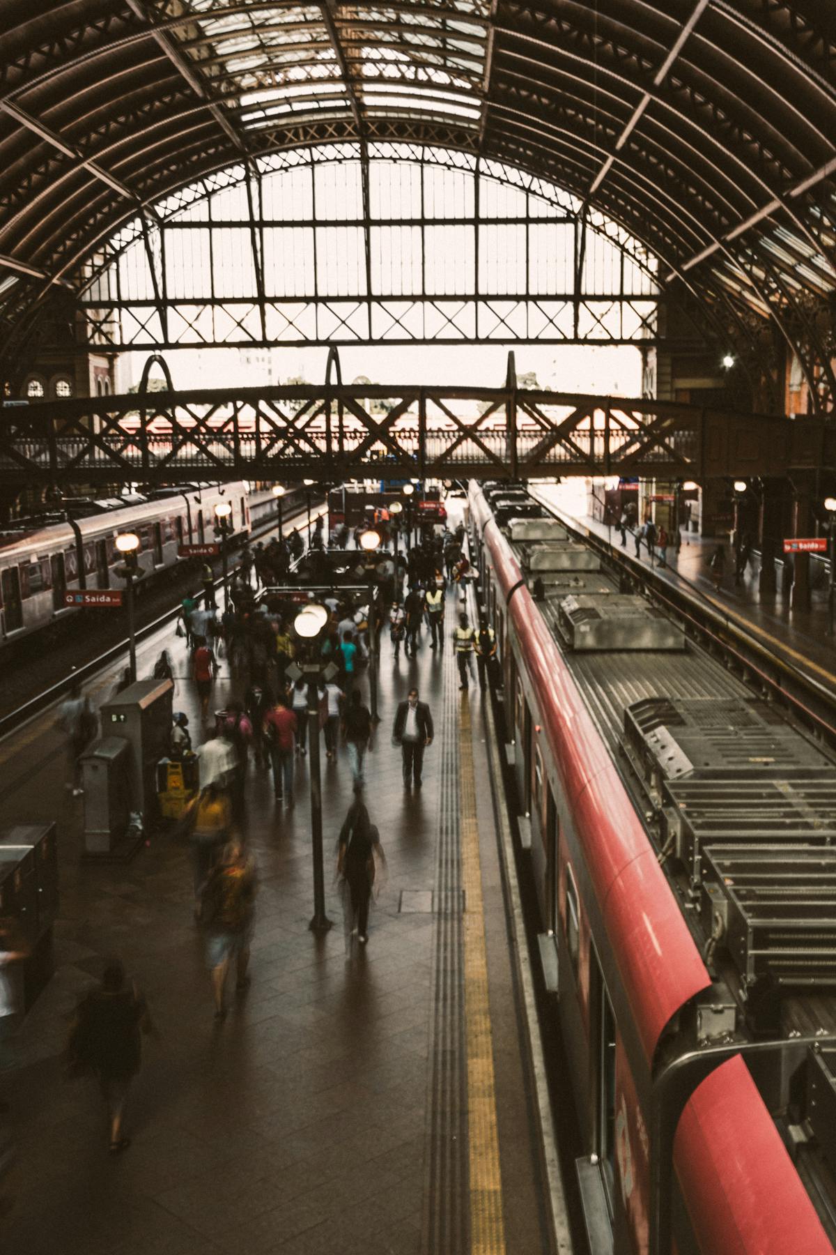 Long covered concourse of an Italian railway station at platform level, with a regional train stopped alongside and commuters walking past