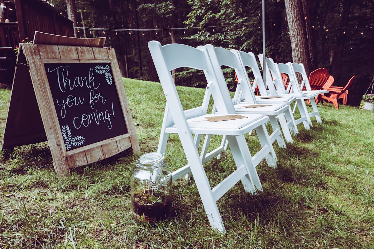 Rows of white folding chairs arranged on grass in front of wooden stage signage, framed by mature trees at the edge of a summer event space
